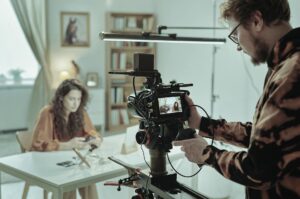 Behind the scenes af en videoproduktion til et E-Learning. Mand bag kamera optager kvinde ved bord.